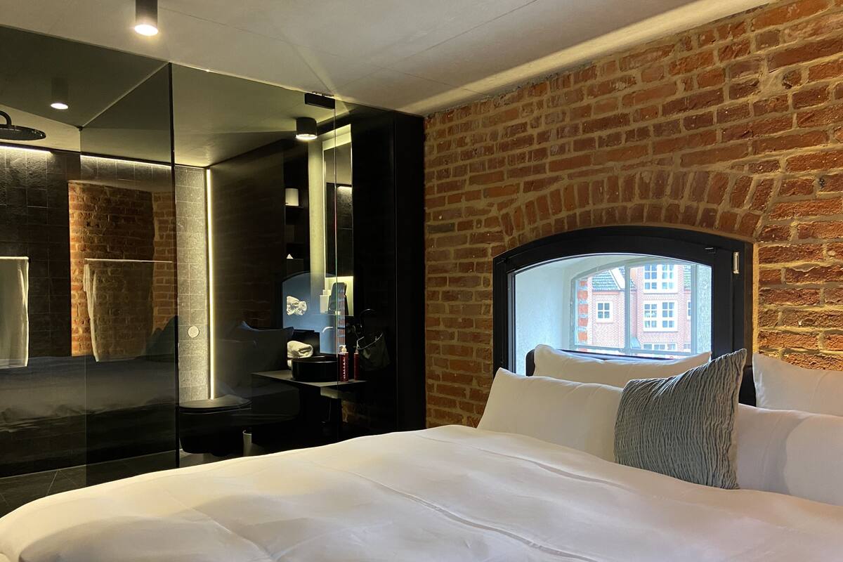 A hotel room with a large bed, a brick wall, a large window, and a mirror.