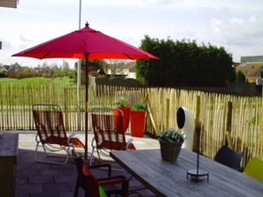 An outdoor patio with a wooden table, two wicker chairs, a red umbrella, and a potted plant. - Nicely furnished house, directly by the sea. With your feet in the sand within 1 minute. (Breskens)