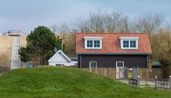 A two-story house with a red-tiled roof, a white fence, and a grassy hill in the foreground. - Nicely furnished house, directly by the sea. With your feet in the sand within 1 minute. (Breskens)