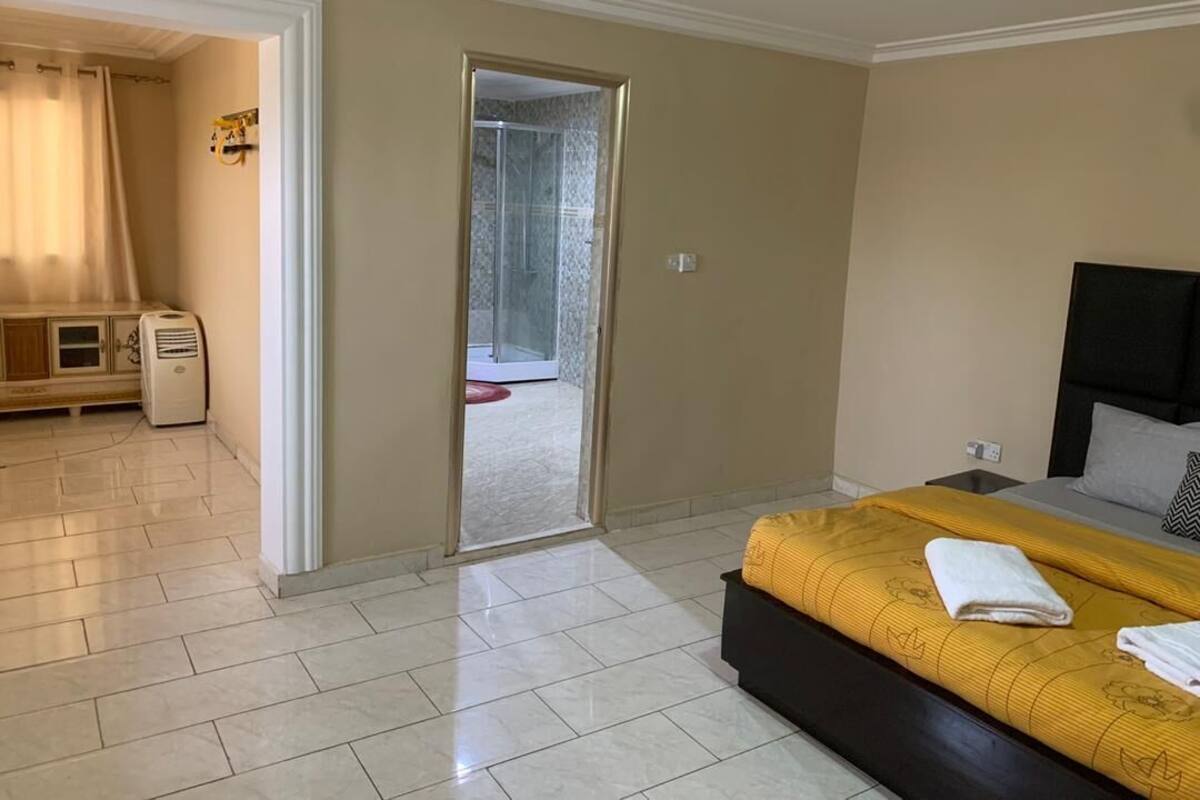 A bedroom with a bed, a nightstand, a bathroom with a shower, and a view of the outside.