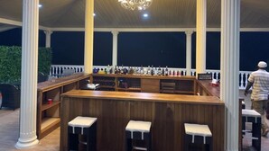 An outdoor bar area with a wooden bar, three stools, and a variety of bottles. - Nimi Beach Resort (Freetown)