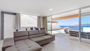 A modern living room with a sectional sofa, a sliding glass door leading to a balcony with a pool and a view of the sea.
