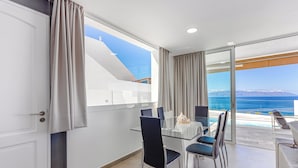 A modern dining area with a glass table and chairs, a view of the sea, and a balcony with a pool.