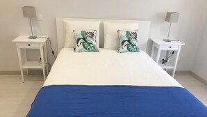 Ein ordentlich bezogenes Bett mit weißer Bettwäsche und zwei Zierkissen, seitlich von zwei Nachttischen mit Lampen und einer Steckdose eingerahmt.