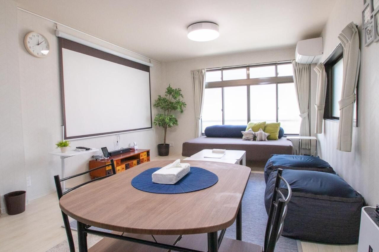 A modern room with a wooden dining table, a sofa, a small desk, a projector screen, and a large window with curtains.