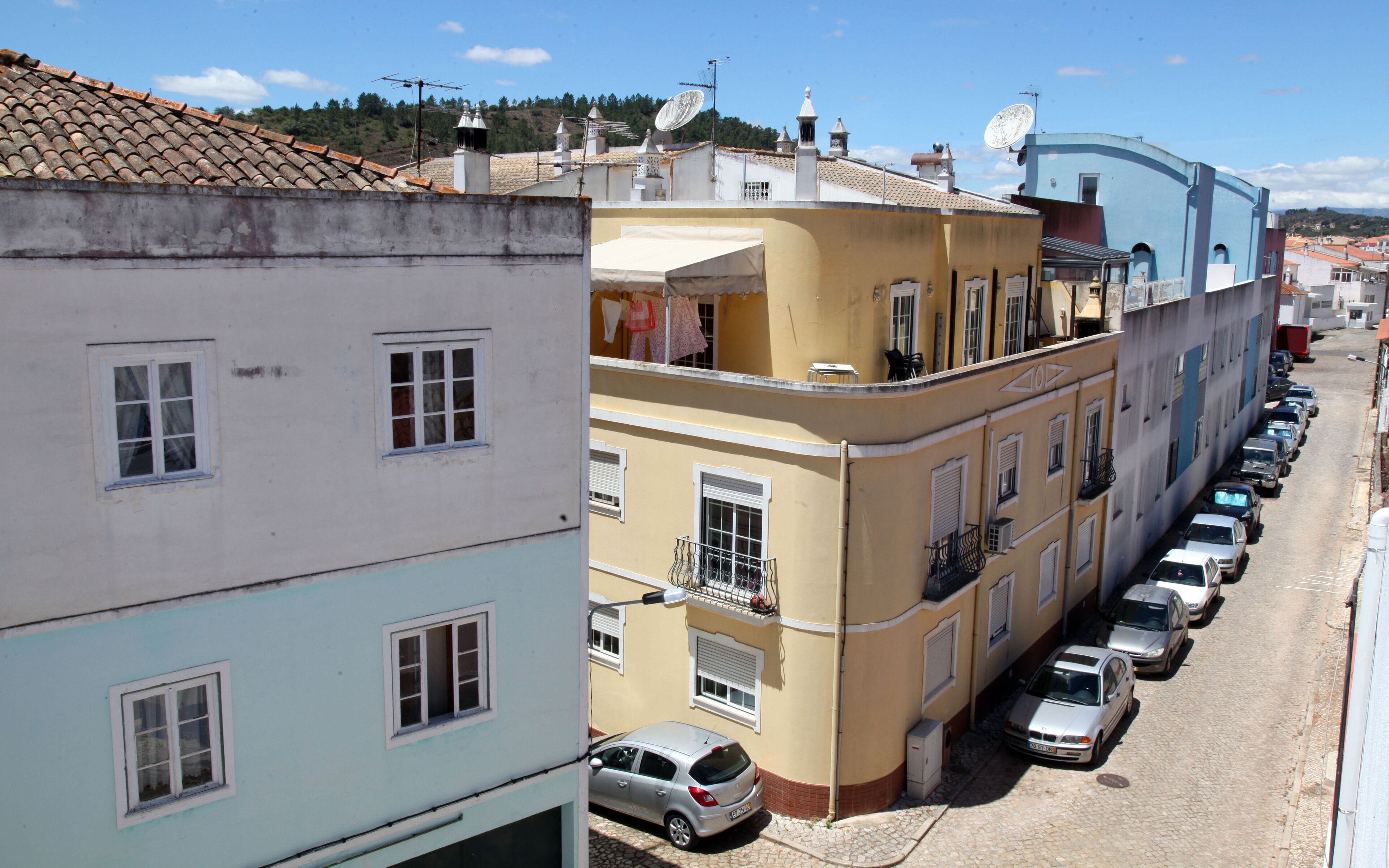 A row of parked cars along a cobblestone street, adjacent to a building with multiple balconies and satellite dishes.