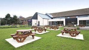 An outdoor area with multiple picnic tables arranged on a grassy lawn, a building with a modern design in the background. - Premier Inn Newbury Town Centre South A339 (Newbury)