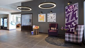 A modern lobby with a reception desk, vending machine, and a seating area with a purple armchair and a plaid armchair. - Premier Inn Newbury Town Centre South A339 (Newbury)