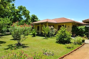 Um edifício amarelo com telhado em tijolos vermelhos, rodeado por um jardim bem cuidado com diversas plantas e flores.
