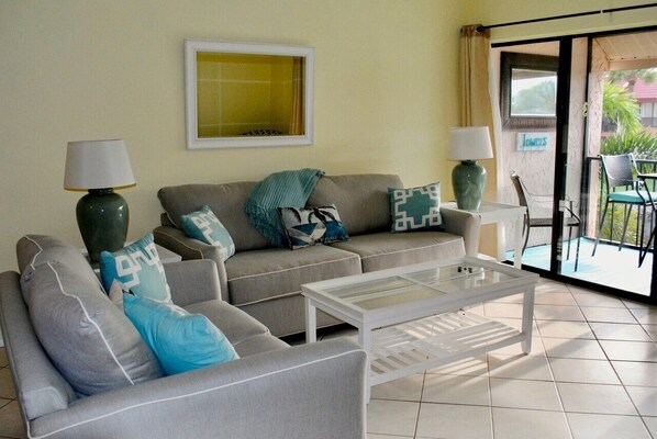 A living room with a sofa set, two side tables with lamps, a glass coffee table, and a sliding glass door leading to an outdoor area.