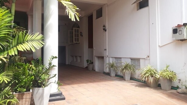 A covered outdoor area with potted plants, a tiled floor, and a white building with columns and windows. - Oasis Inn (Negombo)