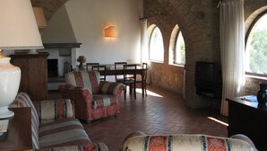 A rustic living room with stone walls, a fireplace, a dining area, and a television. - Agriturismo Chieteno (Cetona)