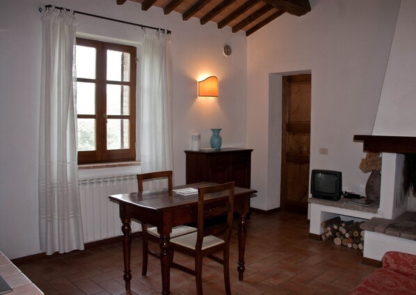 A dining area with a wooden table and chairs, a window with curtains, a wall-mounted lamp, a television, and a fireplace with logs. - Agriturismo Chieteno (Cetona)