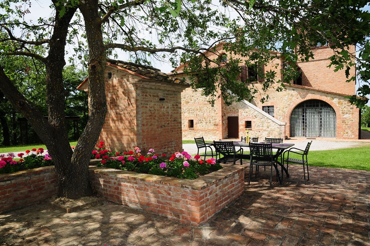Un edificio in mattoni con un patio attrezzato con tavolo e sedie, circondato da aiuole fiorite e un albero.