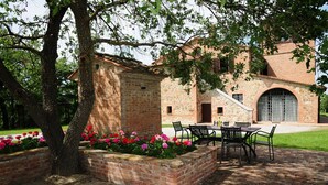 A brick building with a patio area featuring a table and chairs, surrounded by flower beds and a tree. - Agriturismo Chieteno (Cetona)