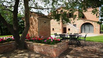 Un edificio in mattoni con un patio attrezzato con tavolo e sedie, circondato da aiuole fiorite e un albero.