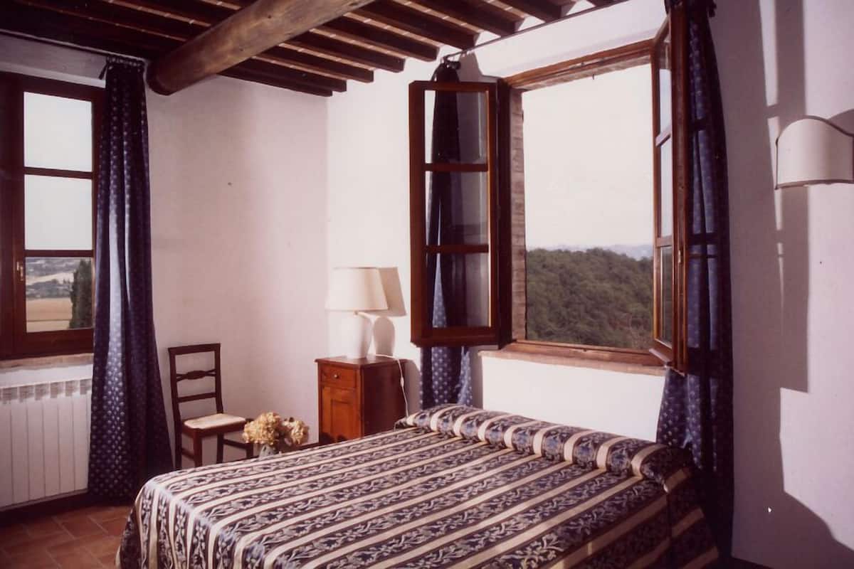 A bedroom with a bed, a chair, a nightstand with a lamp, and two windows with curtains.