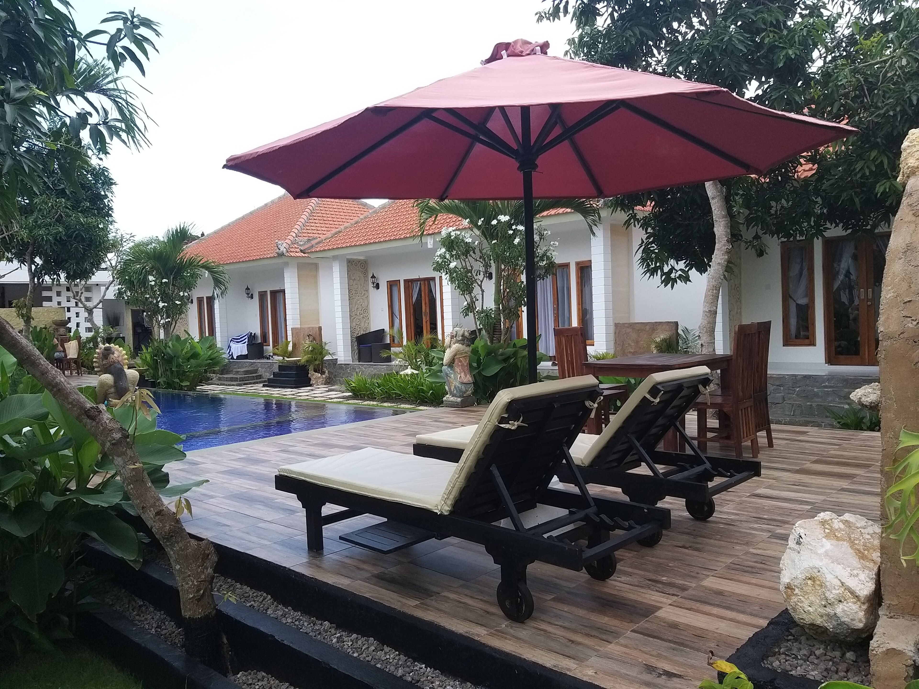 A pool area with lounge chairs and a large umbrella, surrounded by tropical plants and a building with a tiled roof.