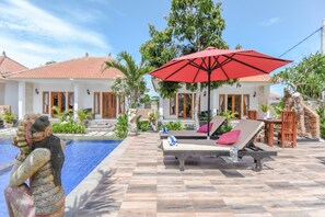 Un área junto a la piscina con sillones de descanso y una sombrilla roja, rodeada por un patio empedrado y follaje.