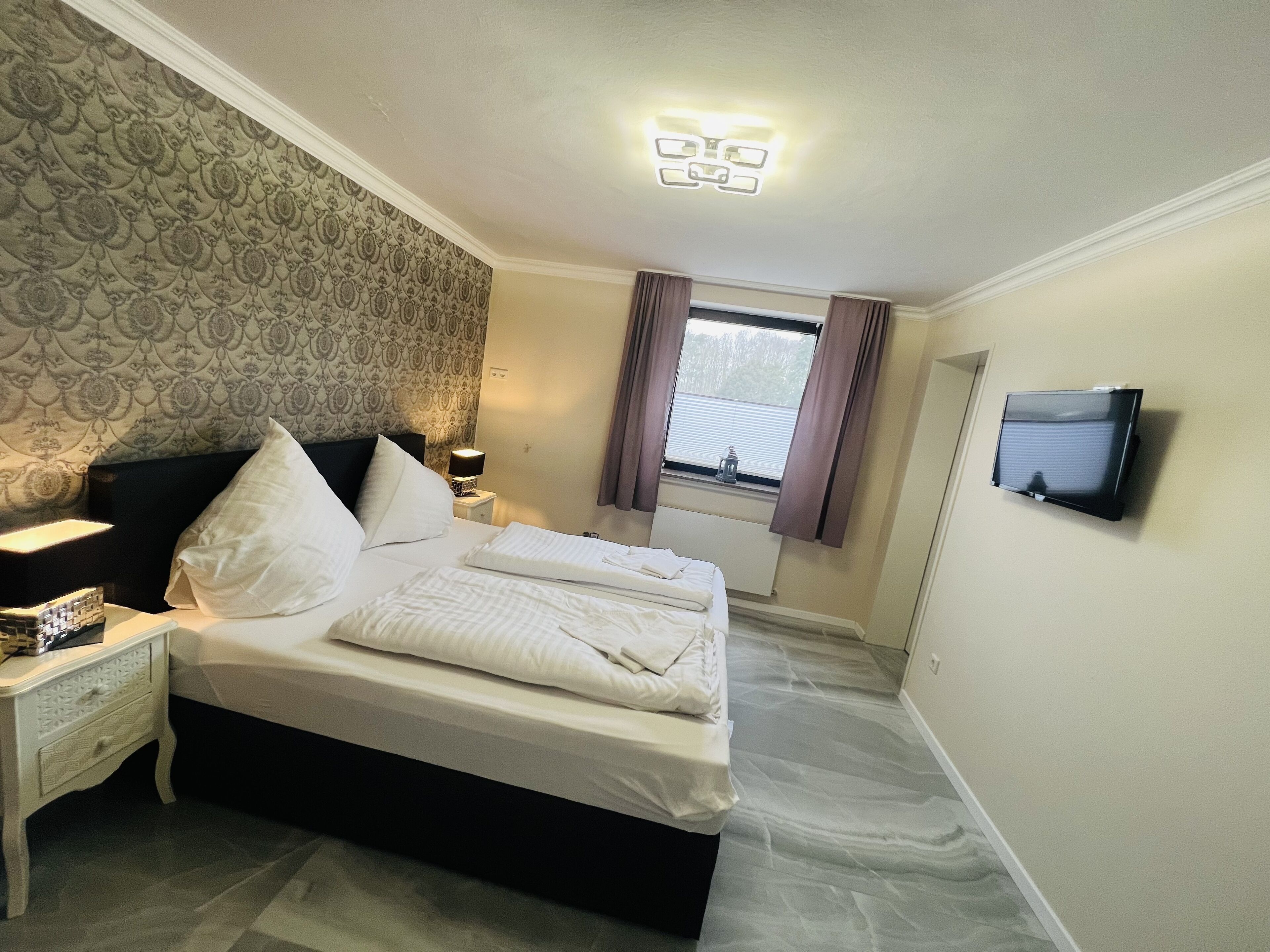 Una cama bien tendida en una habitación de hotel, con papel tapiz estampado, una mesita de noche con lámpara, una ventana con cortinas y un televisor de pantalla plana montado en la pared.