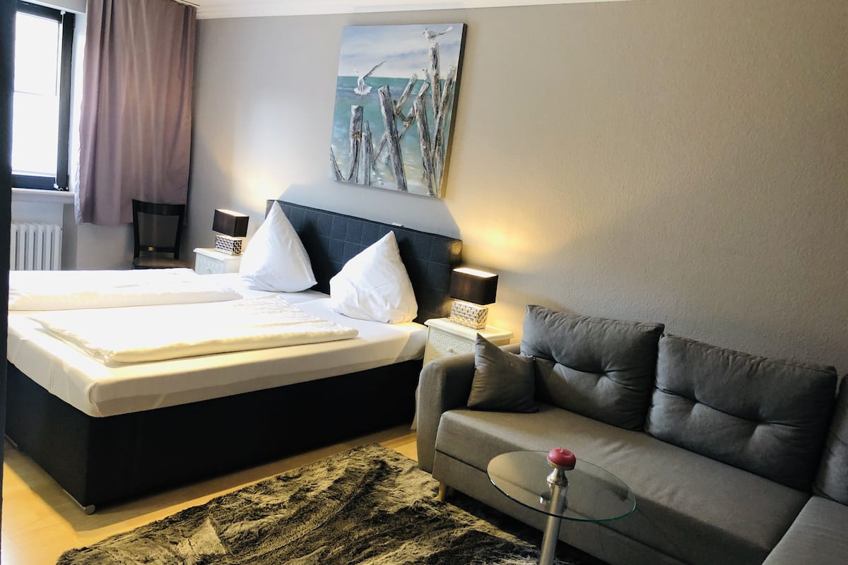 Une chambre d’hôtel avec un lit, un canapé, une table en verre, un tableau au mur et une fenêtre avec des rideaux.