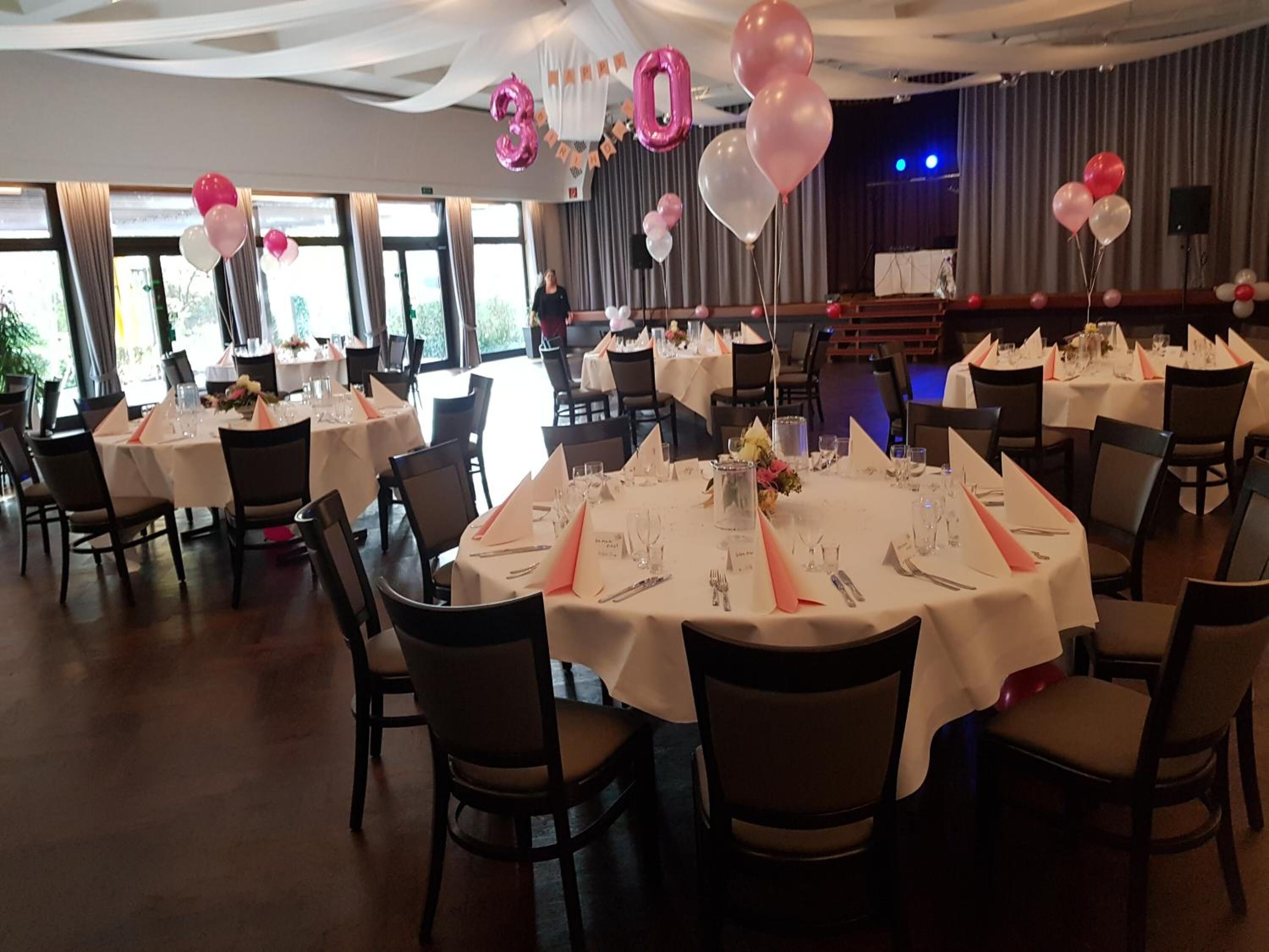 Uma sala de banquetes com mesas redondas preparadas para refeições, decorada com balões rosa e brancos, e um palco com cortinas ao fundo.
