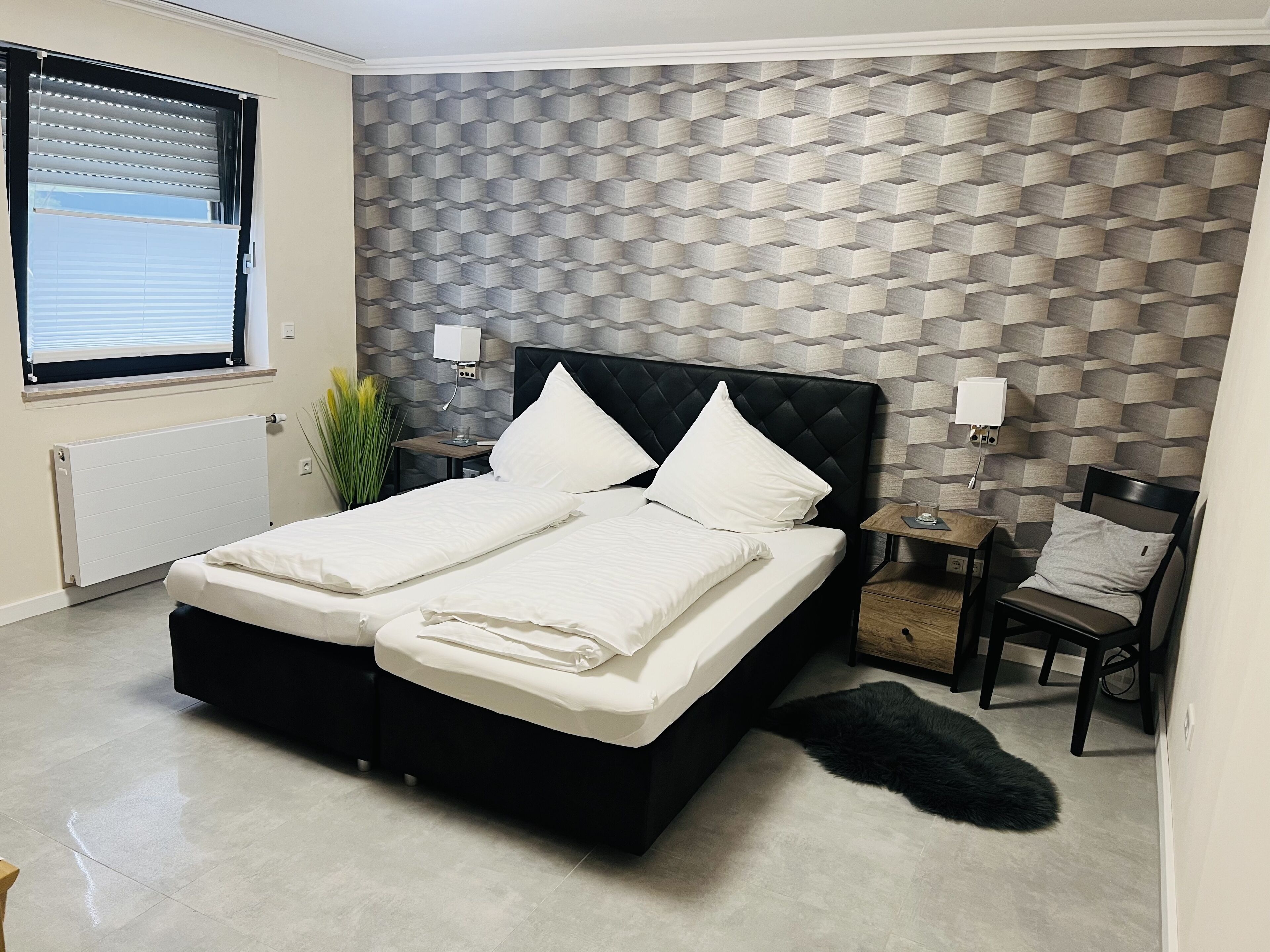 Une chambre moderne avec une tête de lit noire, deux lits blancs, un mur orné d’un motif géométrique, un radiateur, une fenêtre avec des stores, une petite table avec une lampe et un tapis noir à poils longs.