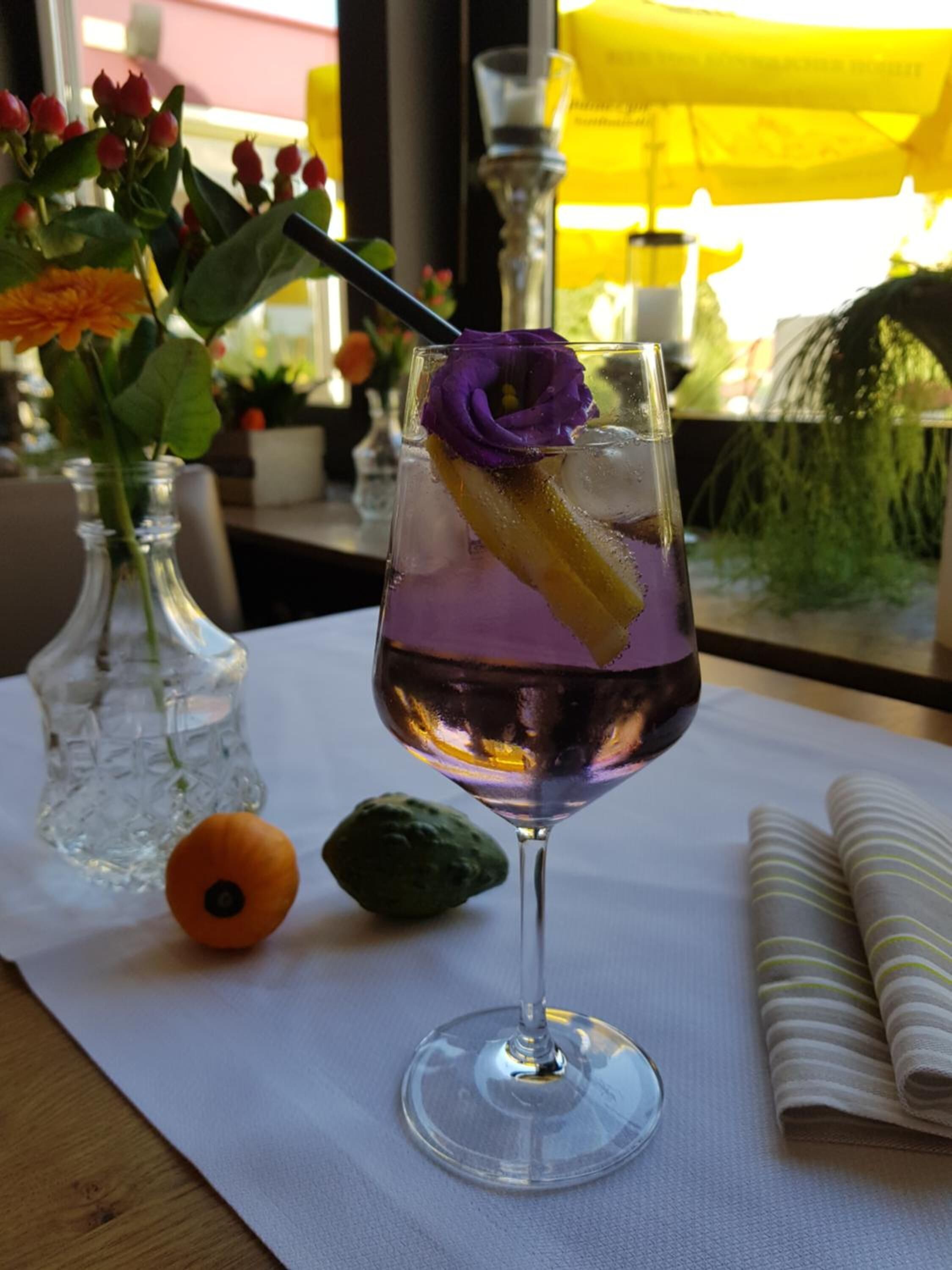 Un verre de cocktail violet, décoré d’une fleur violette, avec des glaçons et une tranche de citron, posé sur une nappe blanche.