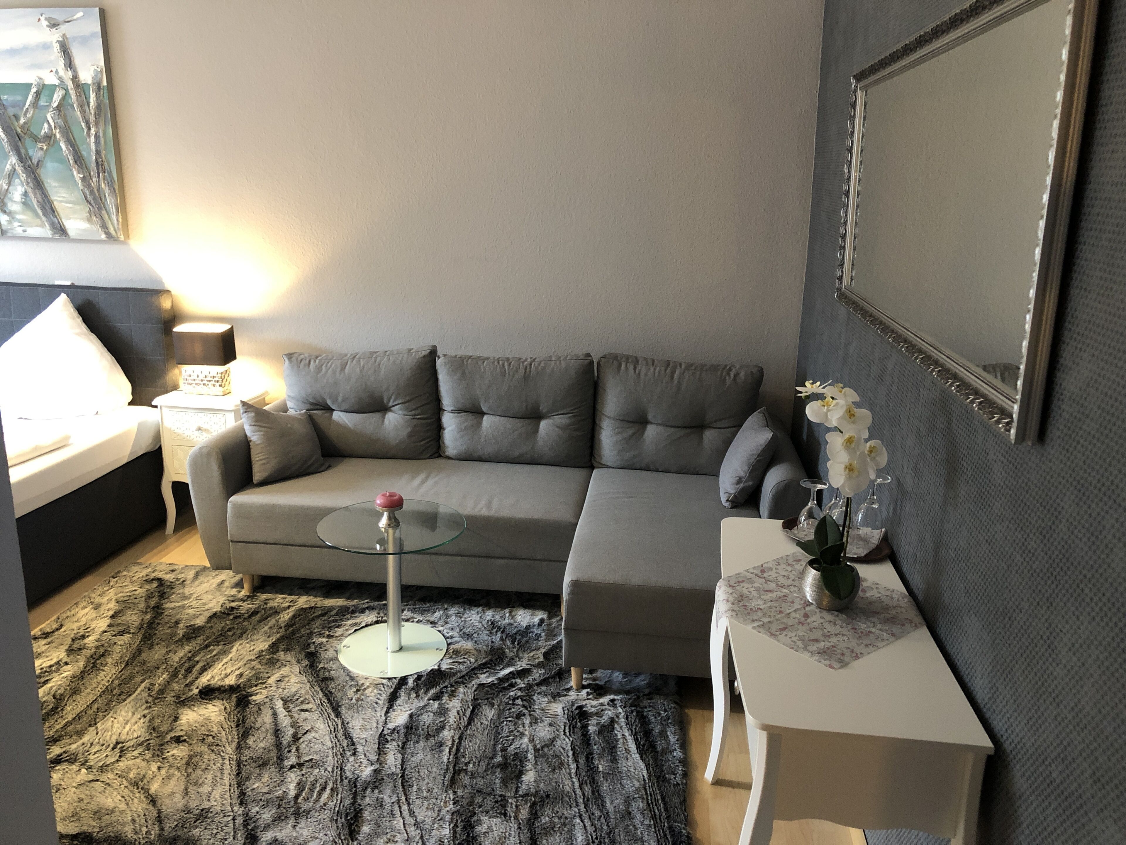 A modern living room with a grey sectional sofa, a glass coffee table, a small side table with a vase of flowers, a lamp, and a painting on the wall.