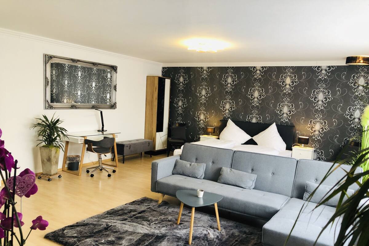 Une chambre moderne avec un canapé gris, un lit avec une literie blanche, un bureau avec une chaise et une grande œuvre d’art encadrée au mur.