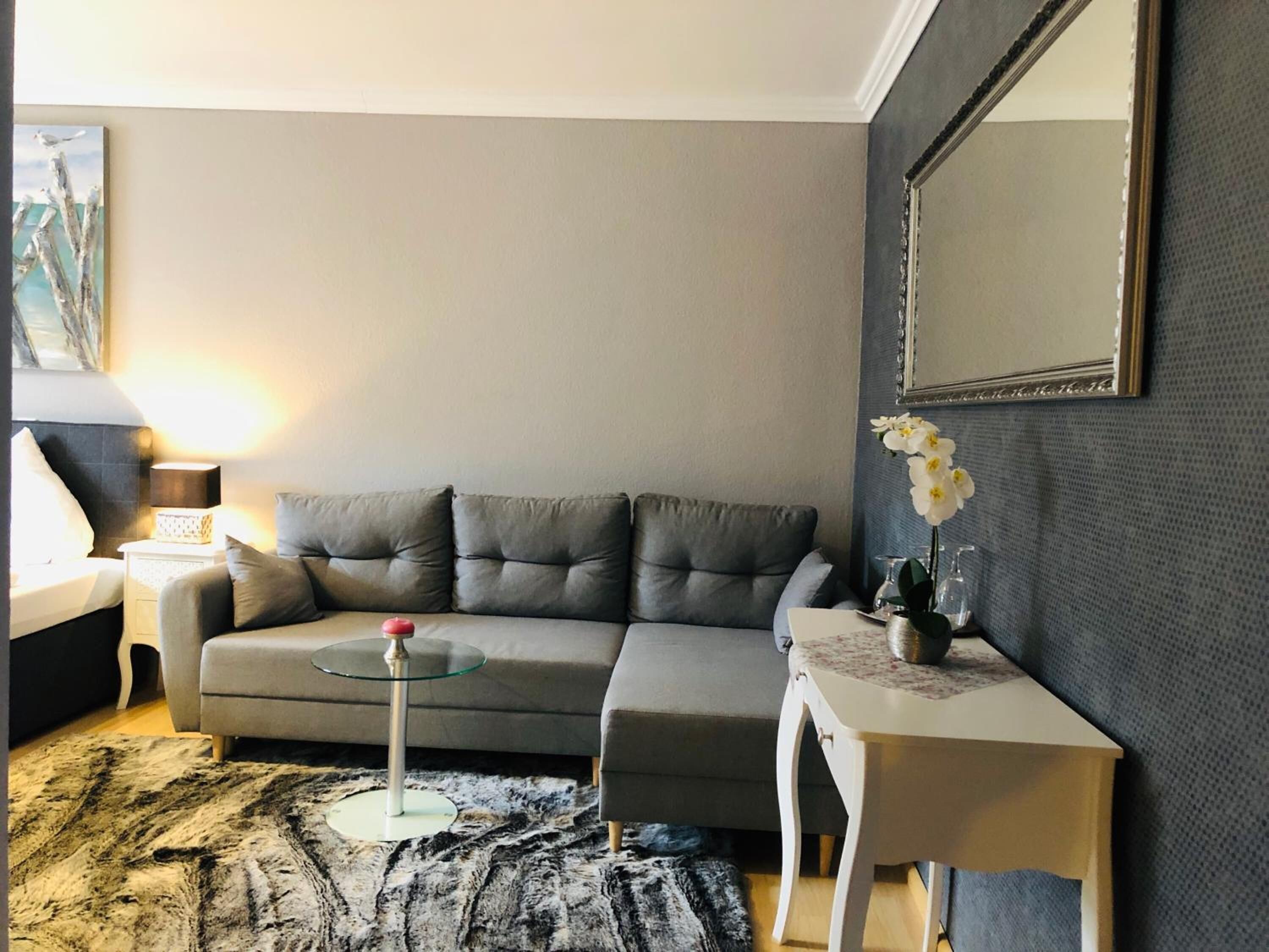 Una sala de estar moderna con un sofá gris, una mesa de centro de vidrio, una mesita auxiliar blanca con un jarrón de flores y un espejo grande en la pared.