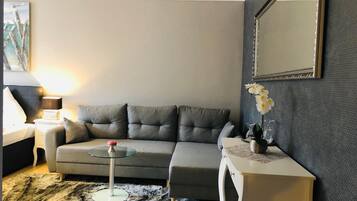 Una sala de estar moderna con un sofá gris, una mesa de centro de vidrio, una mesita auxiliar blanca con un jarrón de flores y un espejo grande en la pared.