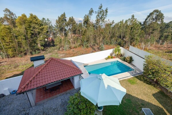 A residential property with a swimming pool, a covered patio, and a view of a forested area.