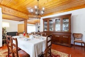A dining room with a wooden ceiling, a chandelier, a dining table set with plates, glasses, and a pitcher. There is a cabinet with glass doors and a chair with a woven seat.