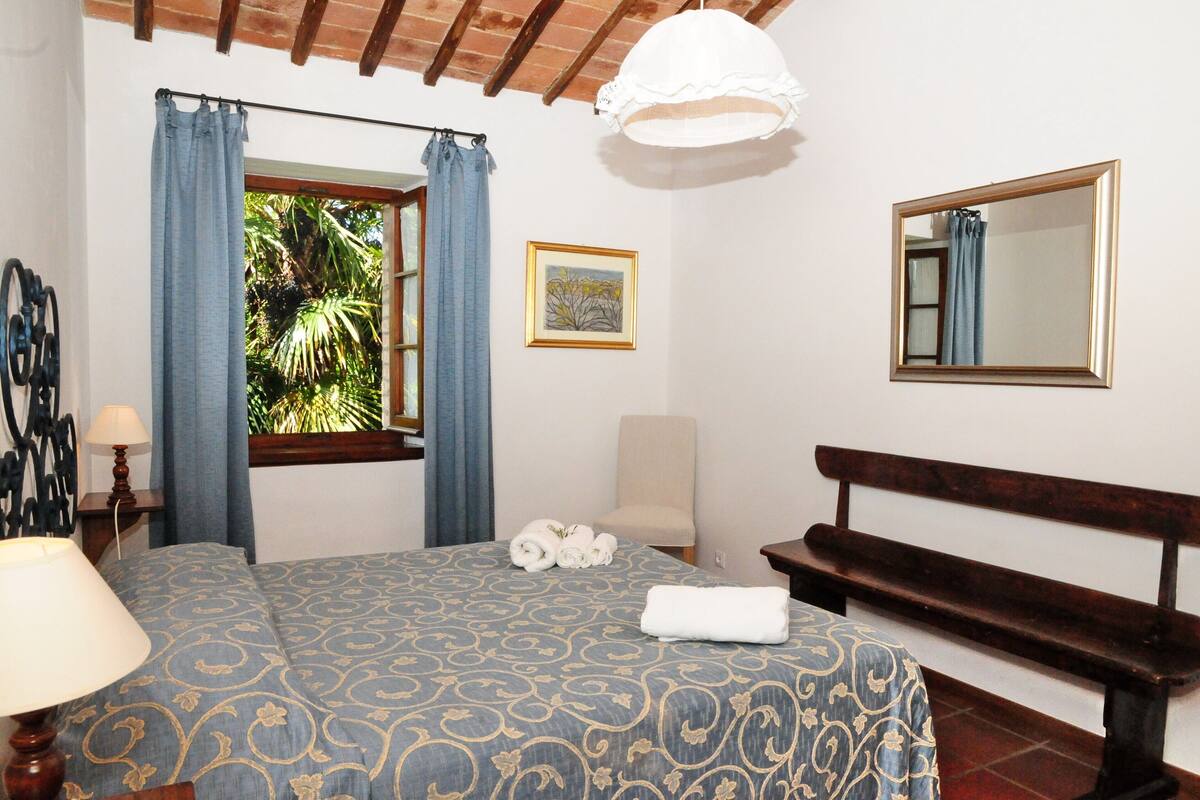 Een slaapkamer met een bed, een bankje, een stoel, een lamp, een raam met gordijnen, een ingelijst kunstwerk en een spiegel.