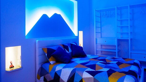 Une chambre à coucher avec une couvre-lit à motifs géométriques, un luminaire fixé au mur et une étagère.