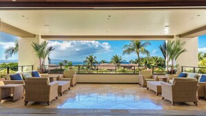 A covered outdoor seating area with wicker furniture and a view of the ocean and palm trees.