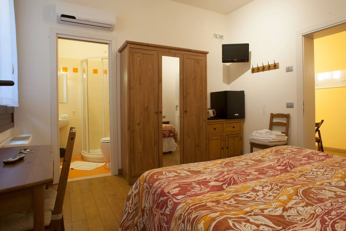 A bedroom with a bed, a wooden wardrobe, a television, a chair, and a bathroom visible through an open door.
