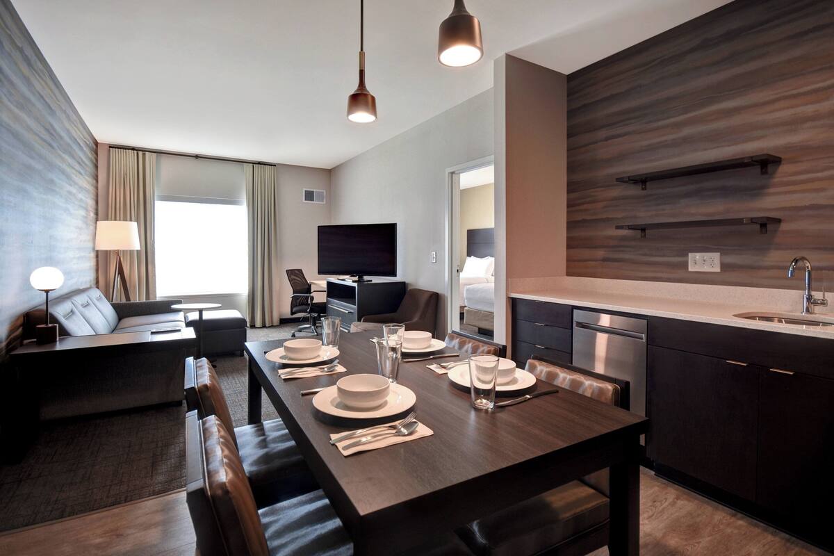 A modern hotel room with a dining area, a sofa, a TV, and a kitchen.