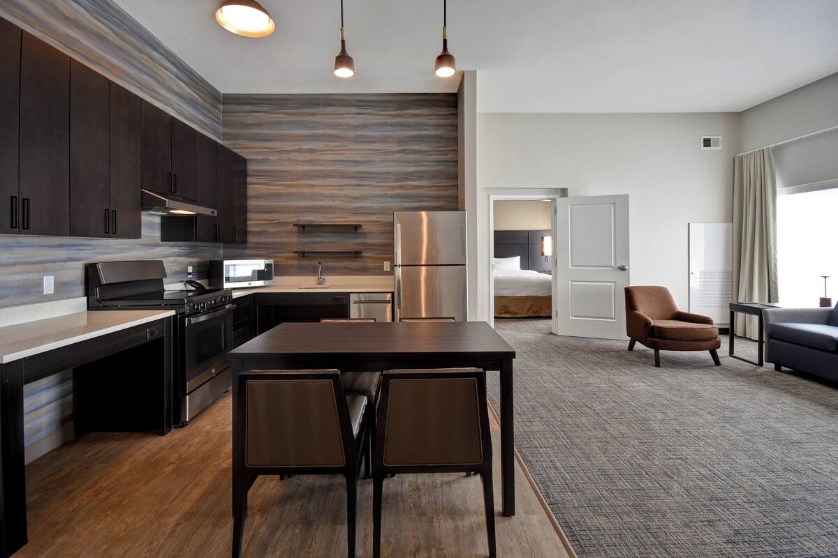 A modern kitchen with dark cabinets, stainless steel appliances, and a wooden backsplash. There is a dining area with a table and chairs, and a living room with a sofa and a chair.
