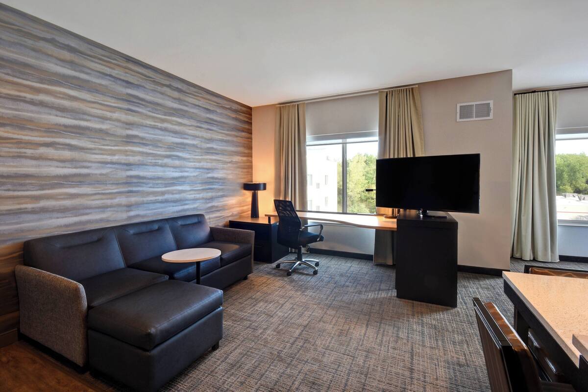 A modern hotel room with a sofa, a small round table, a desk with a chair, a television, and large windows with curtains.