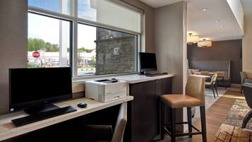 Quarto de hotel moderno com escrivaninha, computador e impressora. Há uma janela grande com vista para o exterior.