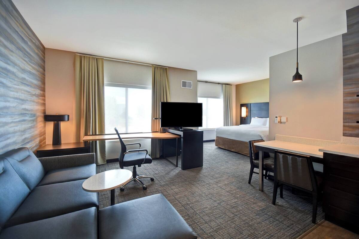 A hotel room with a sofa, a desk with a chair, a bed, a TV, and a window with curtains.