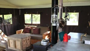 A cozy cabin interior with a wooden sofa, a small table, a stove, and a window with a view of greenery. - Domaine Bonneblond - Tentes Glamping (Saint-Désiré)