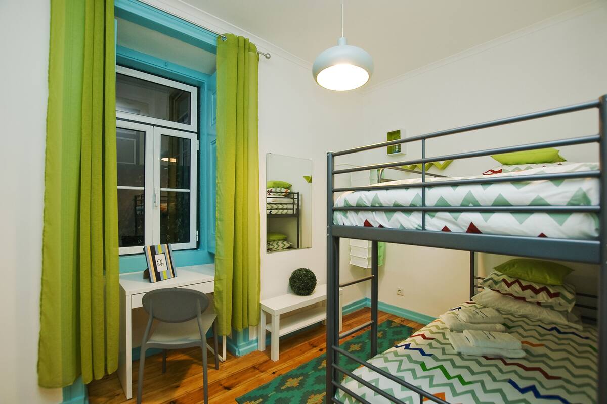 Una cama litera con una colcha verde y blanca de estampado en zigzag, un escritorio con silla, un estante blanco pequeño y una ventana con cortinas verdes.
