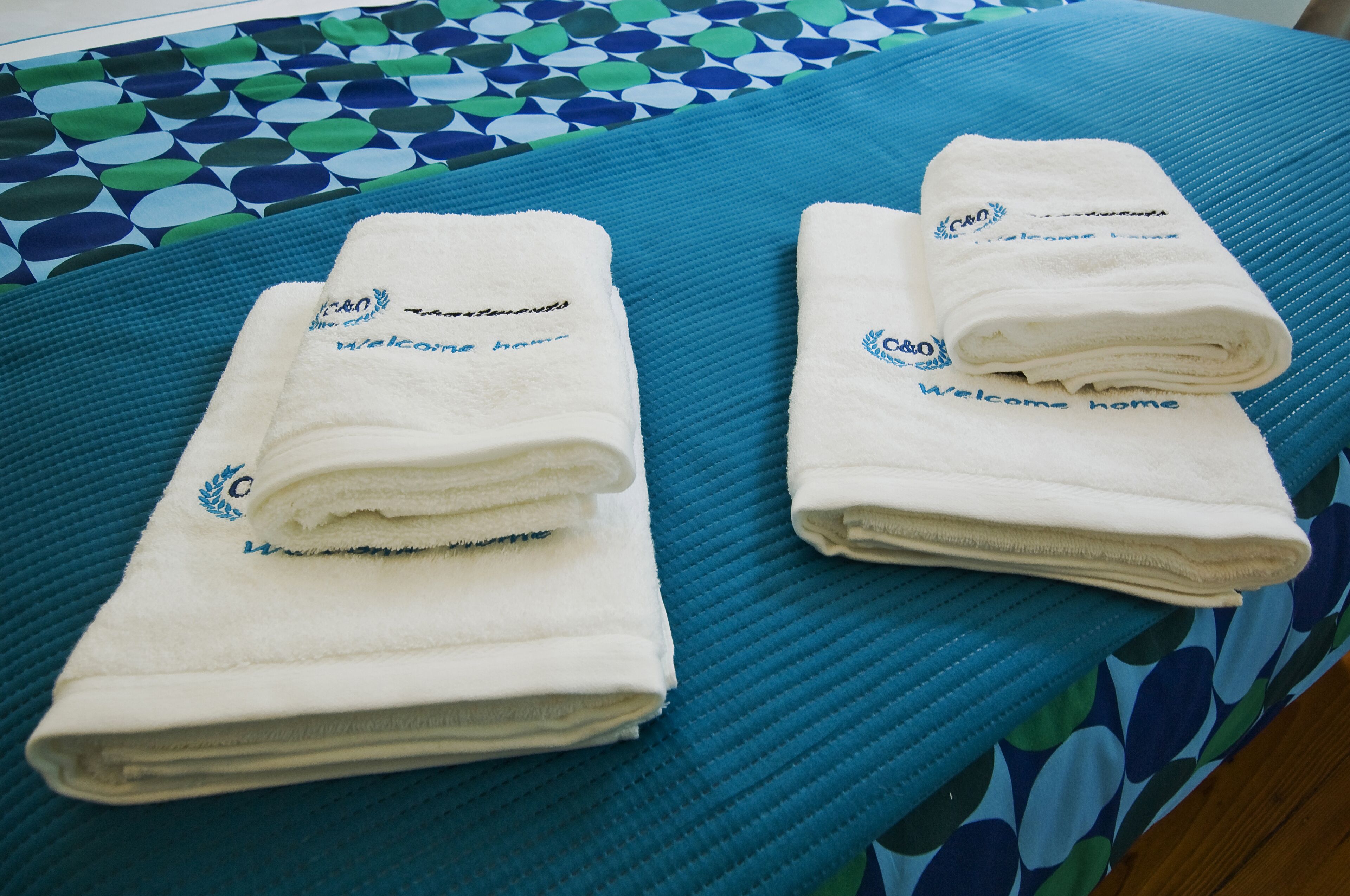 Zwei ordentlich gefaltete weiße Handtücher mit der Aufschrift "Welcome home", auf einer blauen, strukturierten Bettdecke drapiert.
