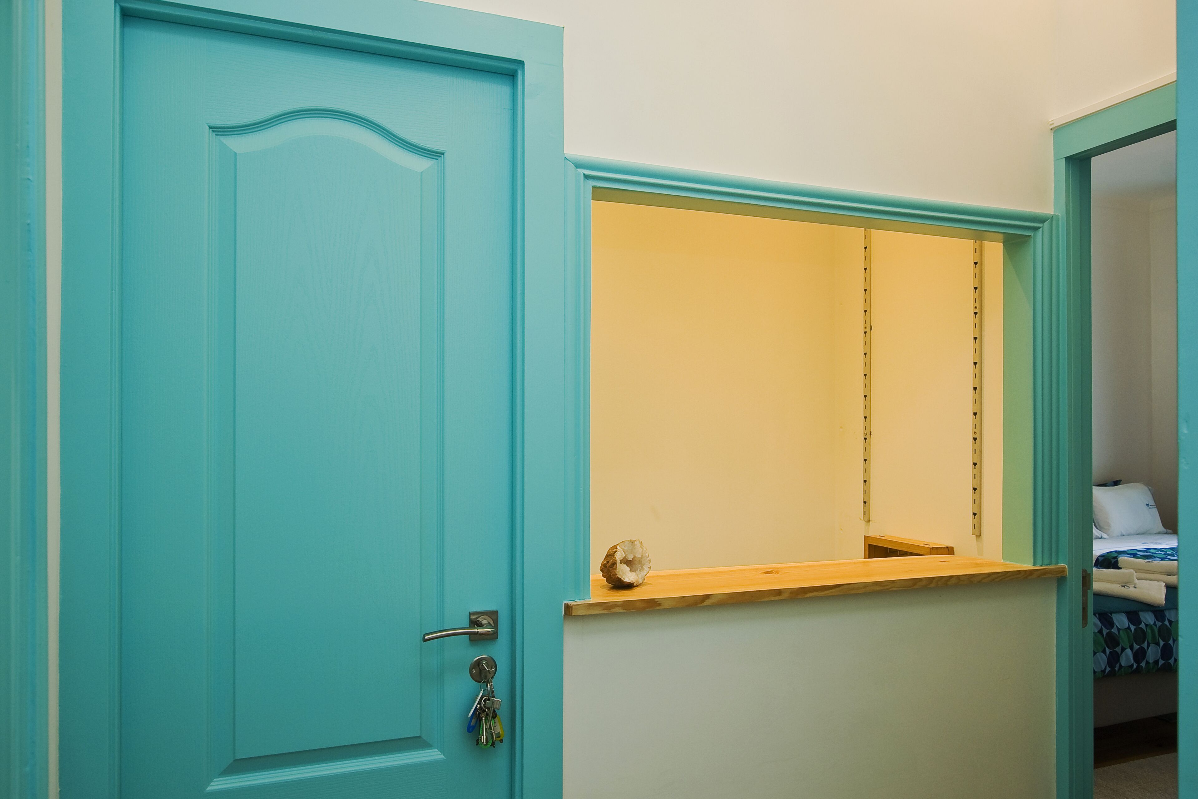 A room with a teal door, a small wooden shelf, and a bed with a blue and white patterned bedspread.