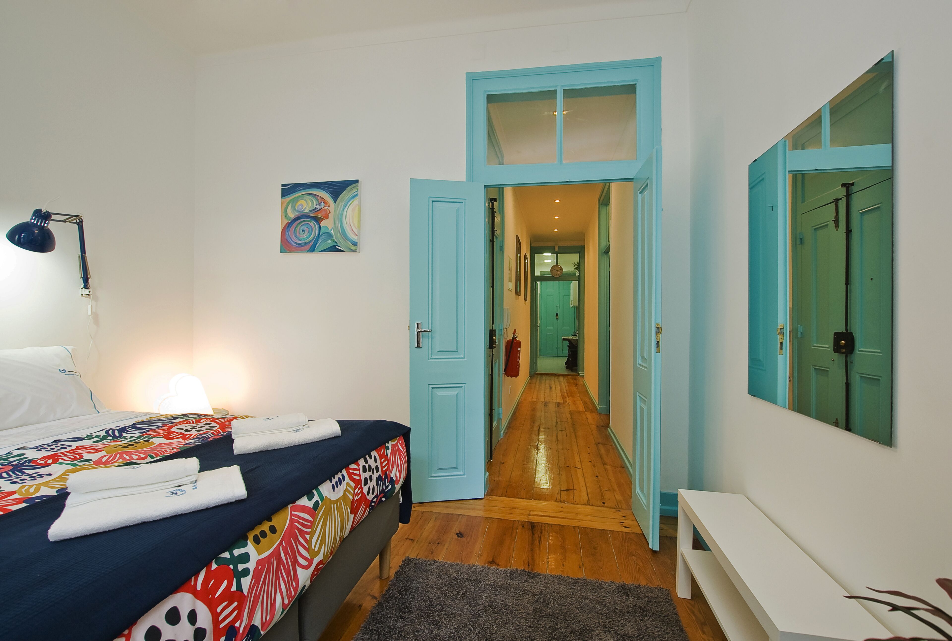 A bedroom with a bed, a desk, a lamp, a painting, and a hallway leading to another room.
