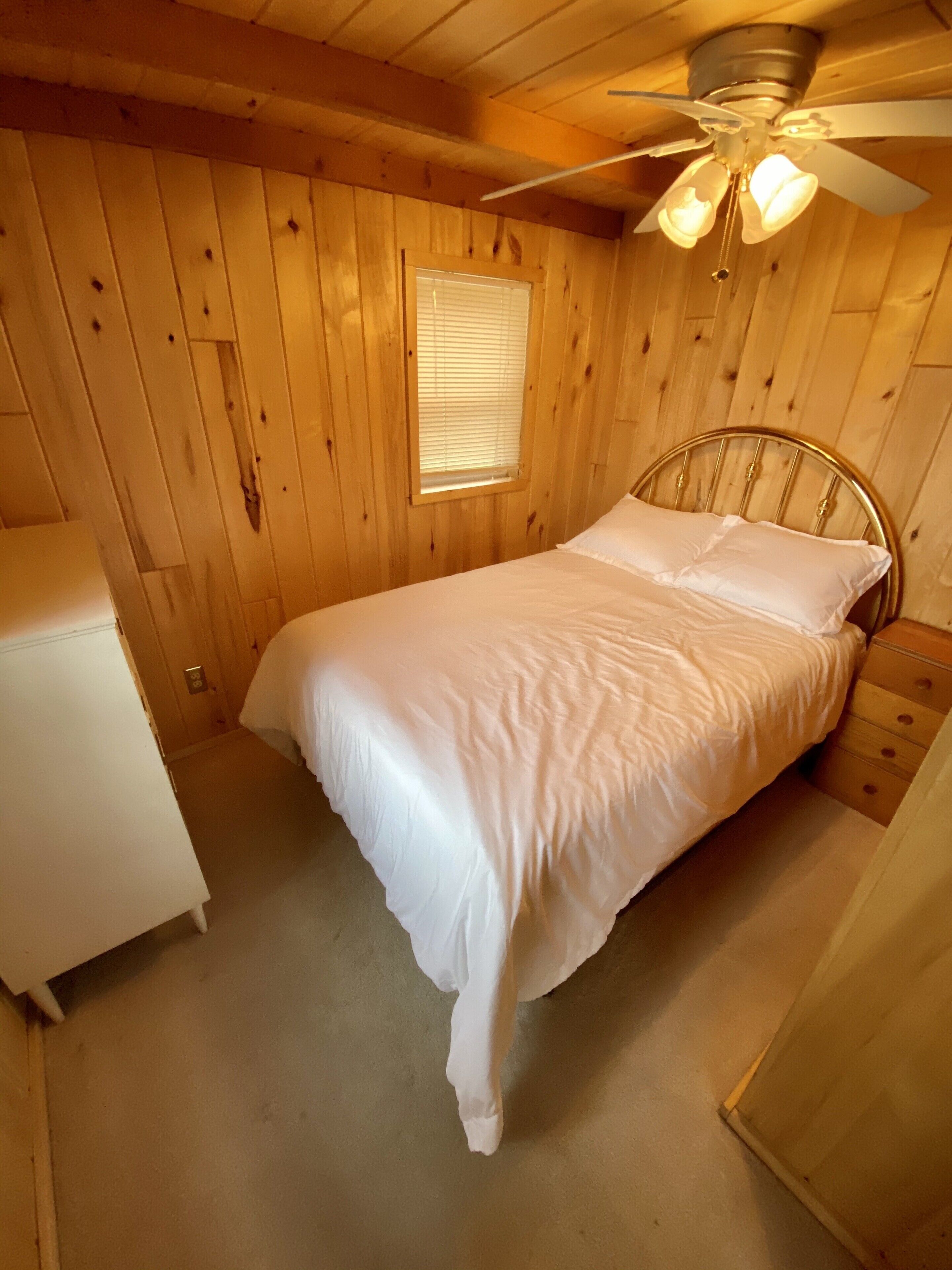 A wooden cabin bedroom with a bed, a ceiling fan, and a window.