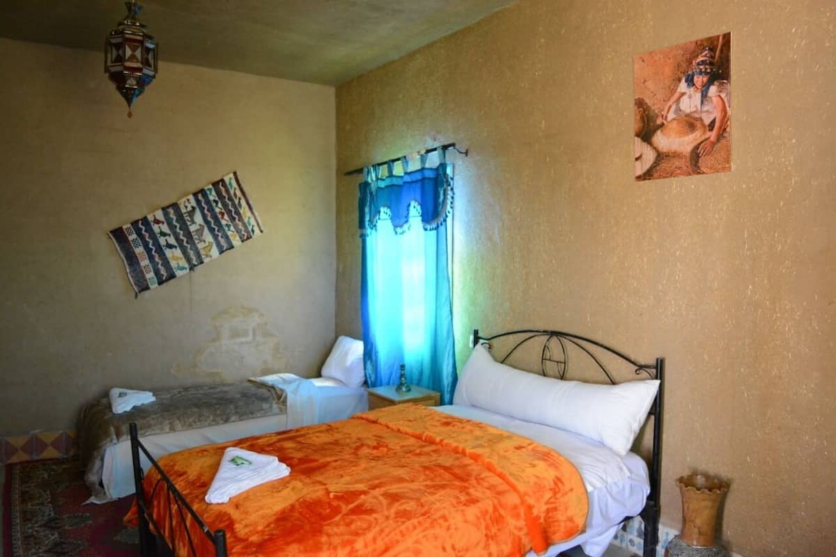 Une chambre à coucher comprenant un lit, une table de chevet, une lampe, une fenêtre avec des rideaux et un tableau ou une décoration murale.